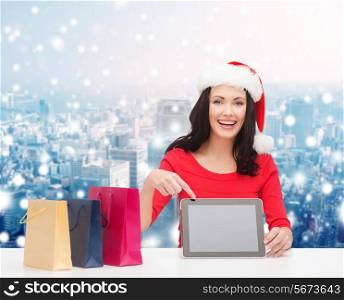 christmas, holidays, technology, advertising and people concept - smiling woman in santa helper hat with shopping bags and tablet pc computer over snowy city background