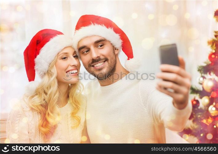 christmas, holidays, technology and people concept - happy couple in santa hats taking selfie picture with smartphone at home over lights