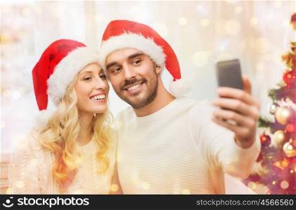 christmas, holidays, technology and people concept - happy couple in santa hats taking selfie picture with smartphone at home over lights