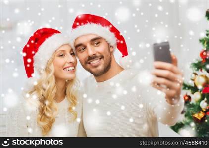 christmas, holidays, technology and people concept - happy couple in santa hats taking selfie picture with smartphone at home