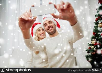 christmas, holidays, technology and people concept - happy couple in santa hats taking selfie picture with smartphone at home