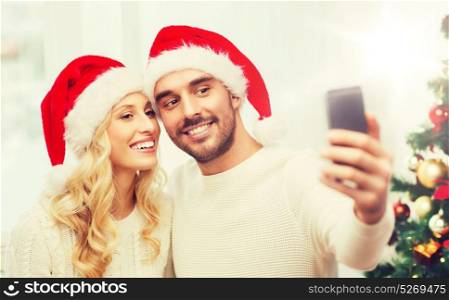 christmas, holidays, technology and people concept - happy couple in santa hats taking selfie picture with smartphone at home. couple taking selfie with smartphone at christmas