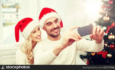 christmas, holidays, technology and people concept - happy couple in santa hats taking selfie picture with smartphone at home. couple taking selfie with smartphone at christmas