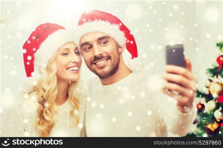 christmas, holidays, technology and people concept - happy couple in santa hats taking selfie picture with smartphone at home. couple taking selfie with smartphone at christmas