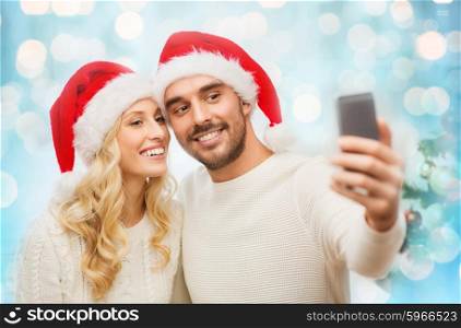 christmas, holidays, technology and people concept - happy couple in santa hats taking selfie picture with smartphone at home over blue lights background