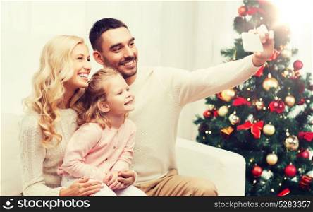 christmas, holidays, technology and people concept - happy family sitting on sofa and taking selfie picture with smartphone at home. family taking selfie with smartphone at christmas