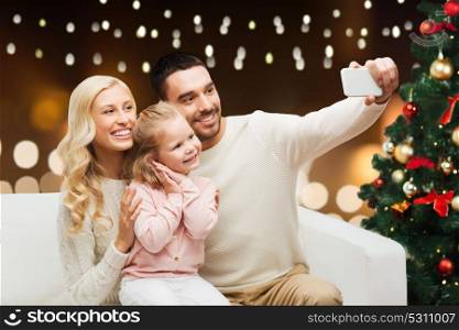christmas, holidays, technology and people concept - happy family sitting on sofa and taking selfie picture with smartphone over lights background. family taking selfie with smartphone at christmas