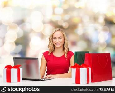 christmas, holidays, technology and people concept - smiling woman in red blank shirt with shopping bags, gifts and laptop computer over lights background