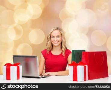 christmas, holidays, technology and people concept - smiling woman in red blank shirt with shopping bags, gifts and laptop computer over beige lights background
