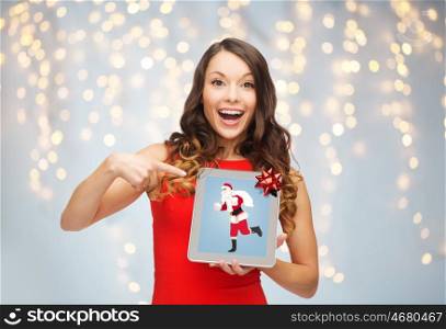 christmas, holidays, technology and people concept - smiling woman in red dress with santa claus picture on tablet pc screen over lights background