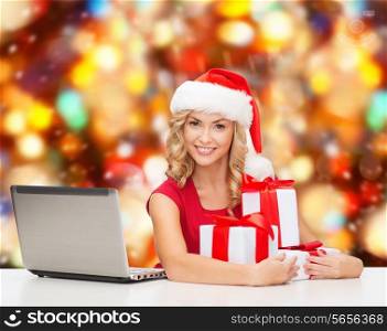 christmas, holidays, technology and people concept - smiling woman in santa helper hat with gifts and laptop computer over red lights background