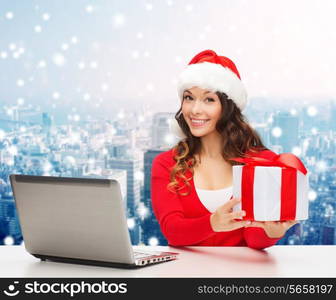 christmas, holidays, technology and people concept - smiling woman in santa helper hat with gift box and laptop computer over snowy city snowing background