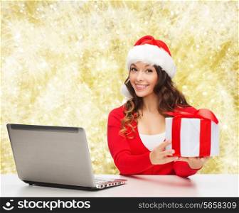 christmas, holidays, technology and people concept - smiling woman in santa helper hat with gift box and laptop computer over yellow snowing background