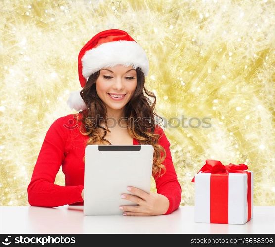 christmas, holidays, technology and people concept - smiling woman in santa helper hat with gift box and tablet pc computer over yellow lights background