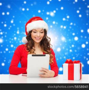 christmas, holidays, technology and people concept - smiling woman in santa helper hat with gift box and tablet pc computer over blue snowy background