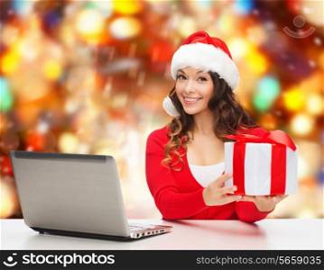christmas, holidays, technology and people concept - smiling woman in santa helper hat with gift box and laptop computer over red snowing background