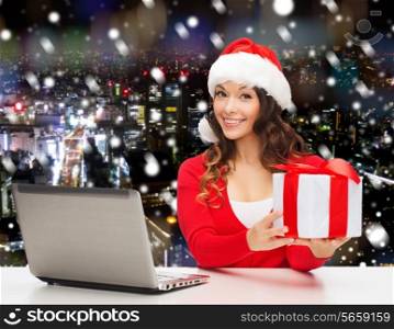 christmas, holidays, technology and people concept - smiling woman in santa helper hat with gift box and laptop computer over snowy night city snowing background