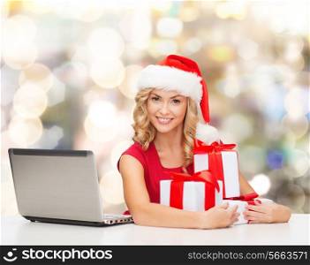 christmas, holidays, technology and people concept - smiling woman in santa helper hat with gifts and laptop computer over lights background