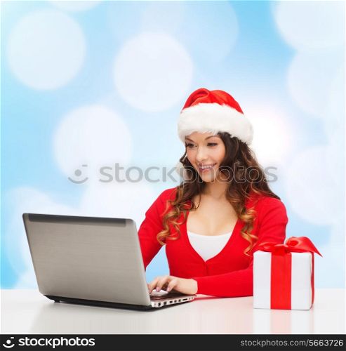 christmas, holidays, technology and people concept - smiling woman in santa helper hat with gift box and laptop computer over blue lights background