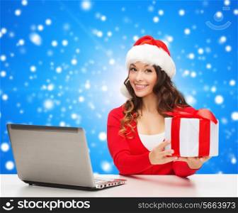 christmas, holidays, technology and people concept - smiling woman in santa helper hat with gift box and laptop computer over blue snowing background