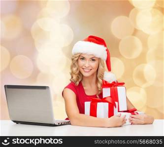 christmas, holidays, technology and people concept - smiling woman in santa helper hat with gifts and laptop computer over beige lights background
