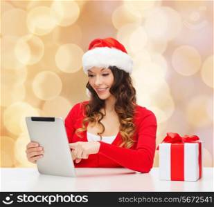christmas, holidays, technology and people concept - smiling woman in santa helper hat with gift box and tablet pc computer over beige lights background