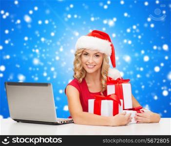 christmas, holidays, technology and people concept - smiling woman in santa helper hat with gifts and laptop computer over blue snowing background