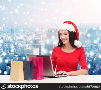 christmas, holidays, technology and people concept - smiling woman in santa helper hat with shopping bags and laptop computer over snowy city background