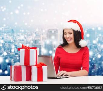 christmas, holidays, technology and people concept - smiling woman in santa helper hat with gift boxes and laptop computer over snowy city background