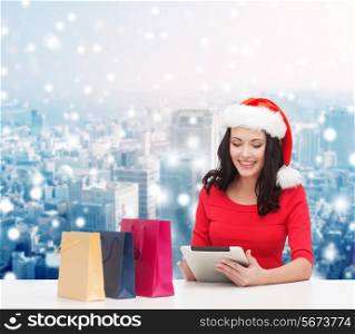 christmas, holidays, technology and people concept - smiling woman in santa helper hat with shopping bags and tablet pc computer over snowy city background