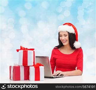 christmas, holidays, technology and people concept - smiling woman in santa helper hat with gift boxes and laptop computer over blue lights background