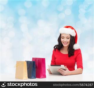 christmas, holidays, technology and people concept - smiling woman in santa helper hat with shopping bags and tablet pc computer over blue lights background