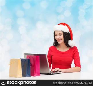 christmas, holidays, technology and people concept - smiling woman in santa helper hat with shopping bags and laptop computer over blue lights background