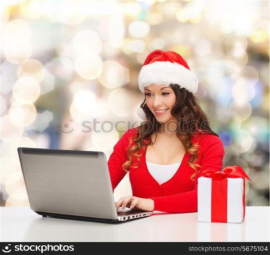 christmas, holidays, technology and people concept - smiling woman in santa helper hat with gift box and laptop computer over lights background