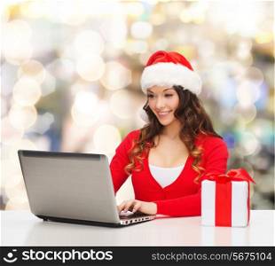 christmas, holidays, technology and people concept - smiling woman in santa helper hat with gift box and laptop computer over lights background