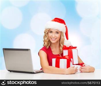 christmas, holidays, technology and people concept - smiling woman in santa helper hat with gifts and laptop computer over blue lights background