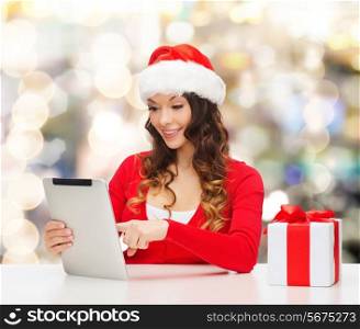 christmas, holidays, technology and people concept - smiling woman in santa helper hat with gift box and tablet pc computer over lights background