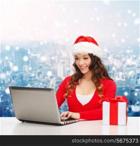 christmas, holidays, technology and people concept - smiling woman in santa helper hat with gift box and laptop computer over snowy city background