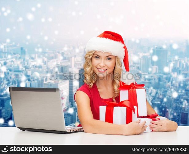 christmas, holidays, technology and people concept - smiling woman in santa helper hat with gifts and laptop computer over snowy city background
