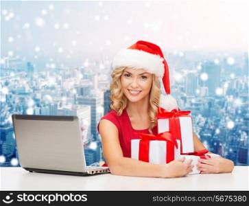 christmas, holidays, technology and people concept - smiling woman in santa helper hat with gifts and laptop computer over snowy city background