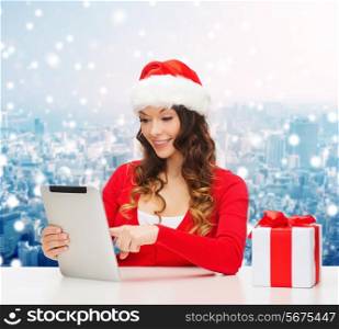 christmas, holidays, technology and people concept - smiling woman in santa helper hat with gift box and tablet pc computer over snowy city background
