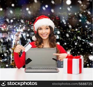 christmas, holidays, technology and people concept - smiling woman in santa helper hat with gift box and laptop computer over snowy night city background
