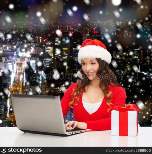 christmas, holidays, technology and people concept - smiling woman in santa helper hat with gift box and laptop computer over snowy night city background