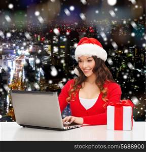 christmas, holidays, technology and people concept - smiling woman in santa helper hat with gift box and laptop computer over snowy night city background