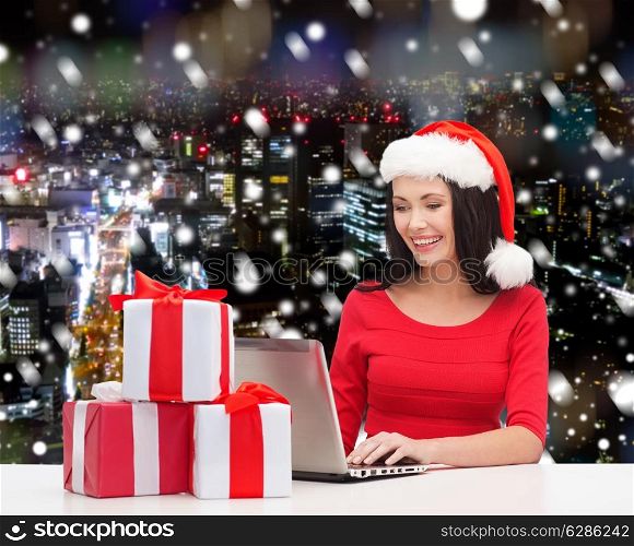 christmas, holidays, technology and people concept - smiling woman in santa helper hat with gift boxes and laptop computer over snowy night city background