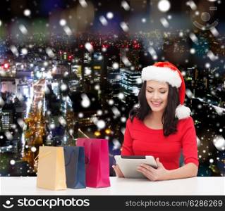 christmas, holidays, technology and people concept - smiling woman in santa helper hat with shopping bags and tablet pc computer over snowy night city background