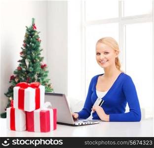 christmas, holidays, technology and shopping concept - smiling woman gift boxes, credit card and laptop computer over living room with christmas tree background