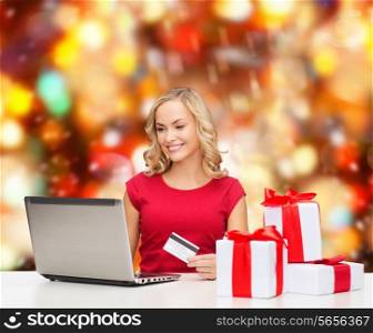 christmas, holidays, technology and shopping concept - smiling woman in red blank shirt with gift boxes, credit card and laptop computer over red lights background