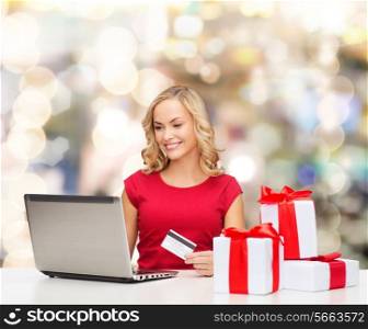 christmas, holidays, technology and shopping concept - smiling woman in red blank shirt with gift boxes, credit card and laptop computer over lights background