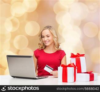 christmas, holidays, technology and shopping concept - smiling woman in red blank shirt with gift boxes, credit card and laptop computer over beige lights background